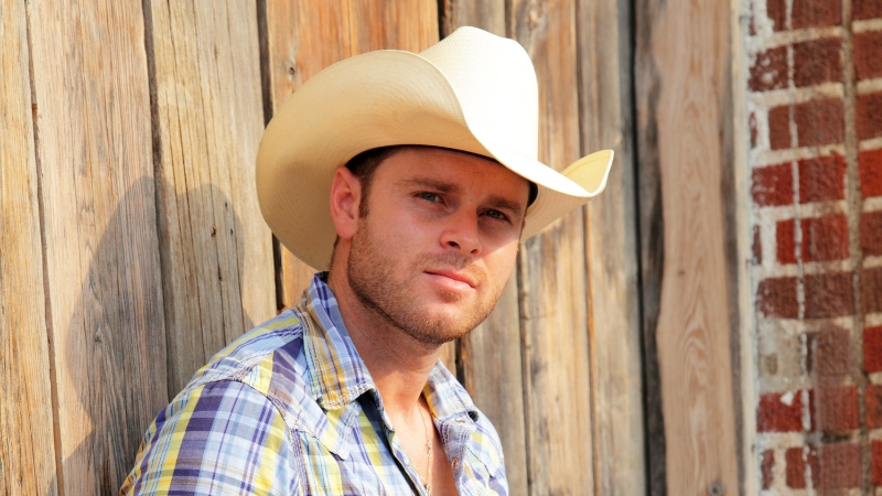 A man in a cowboy hat and plaid shirt stands by a wooden wall