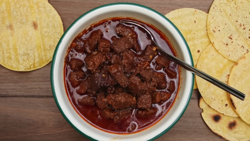 A bowl of authentic Texas chili with chunks of beef in a rich red sauce, served with corn tortillas on a wooden table