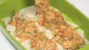 A spoonful of chicken and vegetable filling is being spread over a layer of torn corn tortillas in a green baking dish