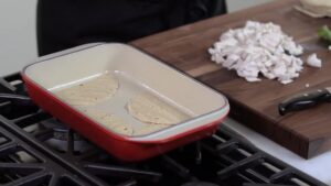 Corn tortillas are being layered in a red baking dish on the stove next to a cutting board with chopped cooked chicken