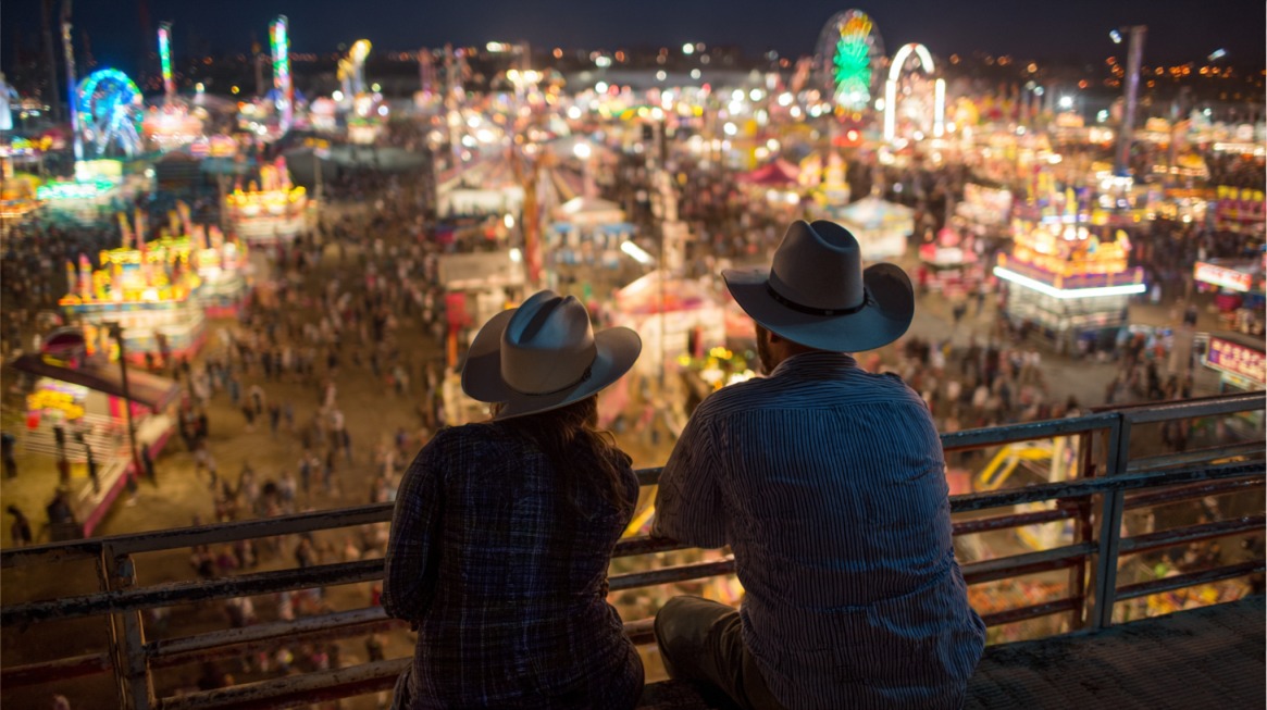 Two people in cowboy hats overlooking a brightly lit fairground at night with rides and crowds